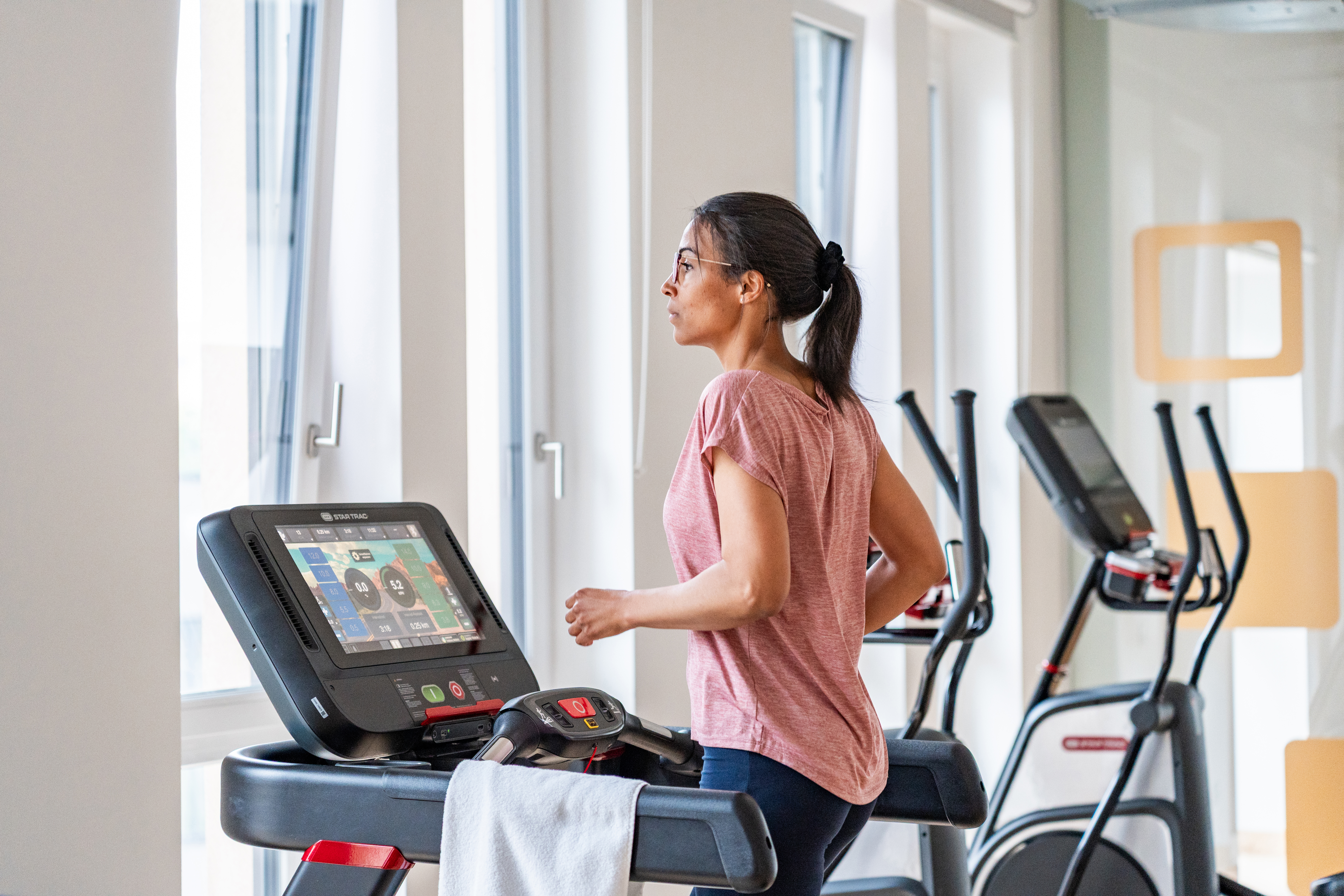 Seitenansicht einer Frau mit rosa Shirt, die mit Blick aus dem Fenster auf einem Hometrainer läuft.