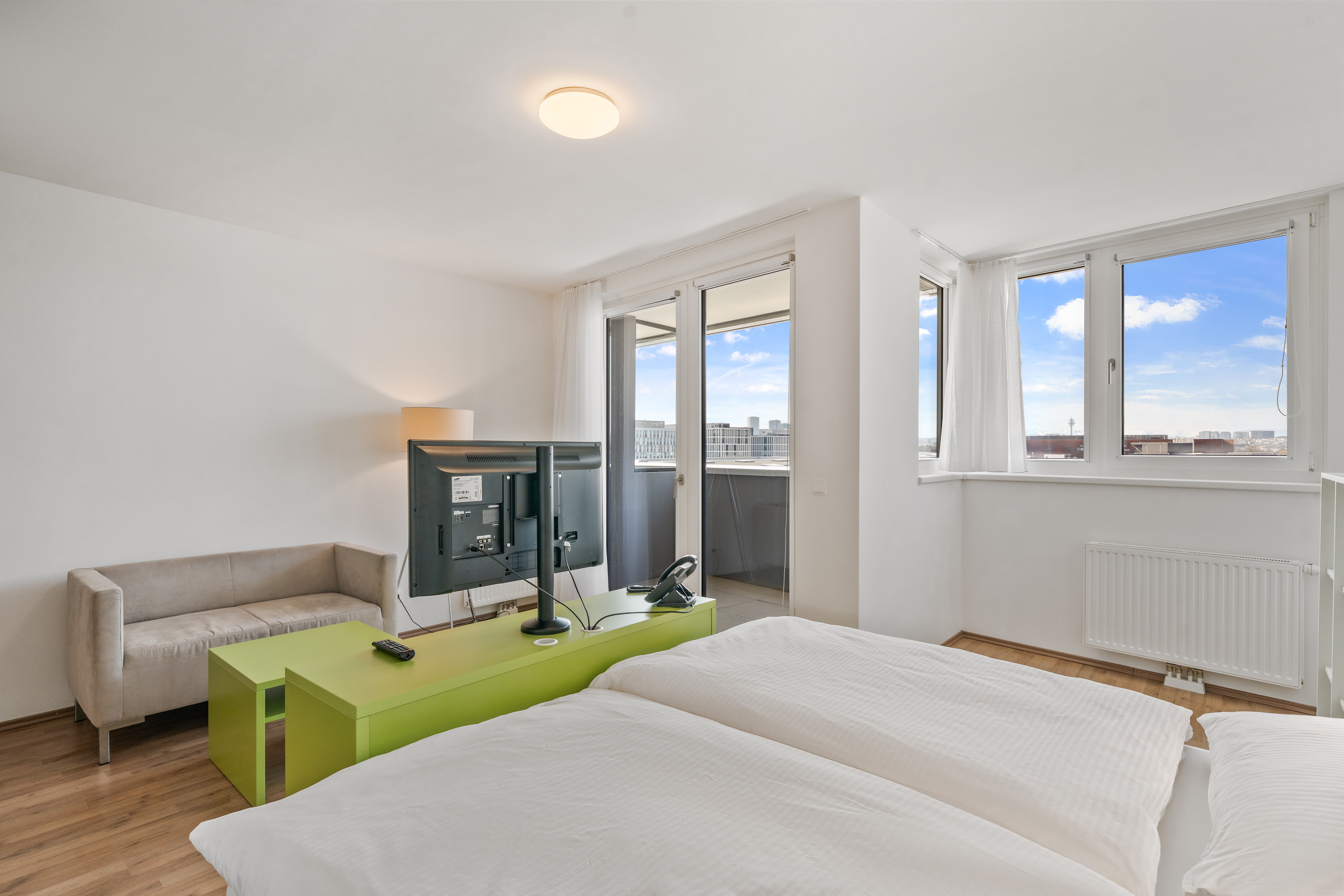 Wohn-Schlafbereich eines möblierten Room4Rent Apartments mit Doppelbett, Arbeitsplatz mit grünem Schreibtisch und großem Balkon mit Stadtblick