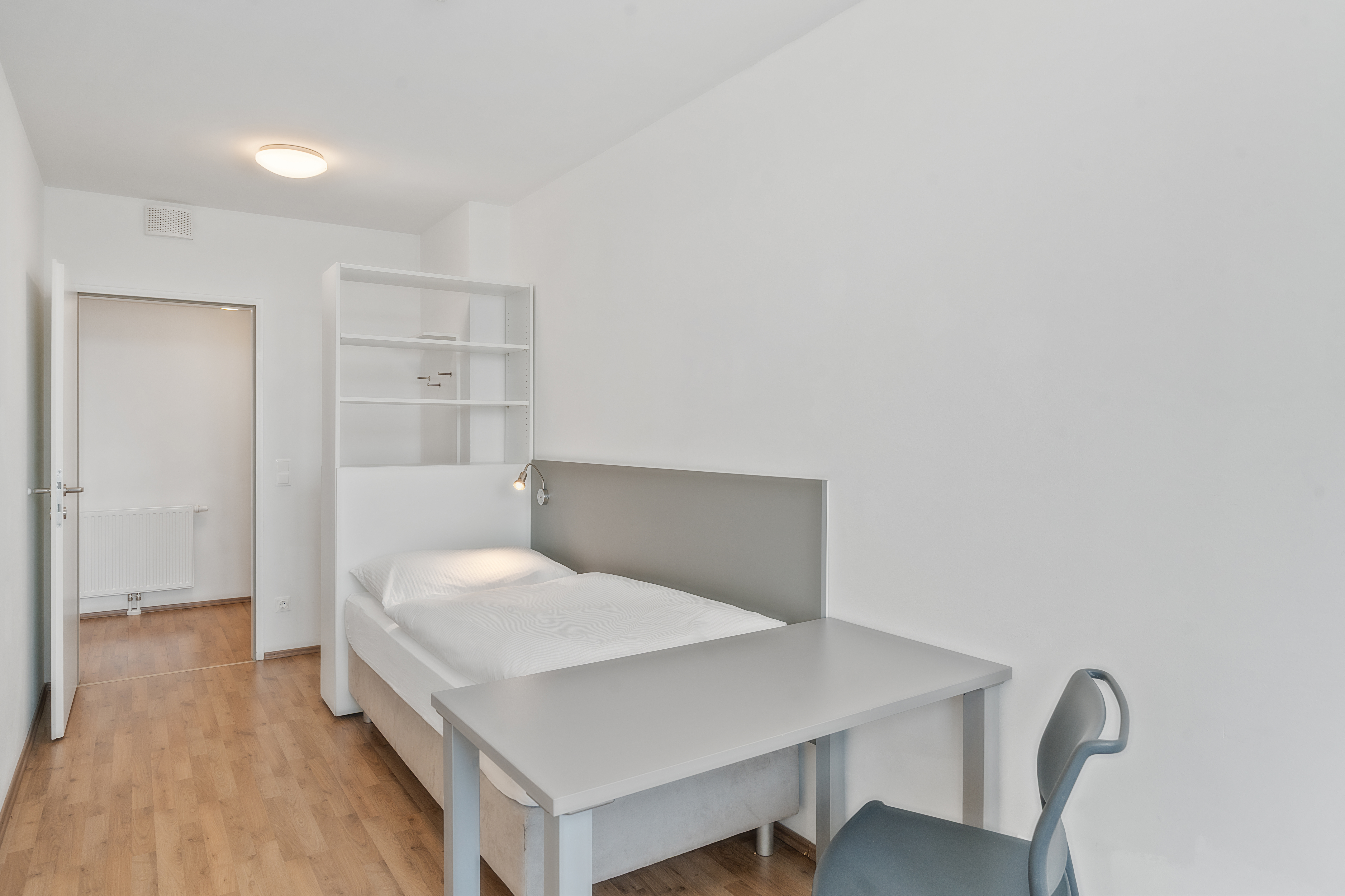 Schlafbereich eines leeren Apartments Typ XL mit Bett, grauem Kopfteil, Arbeitstisch und Wandregal