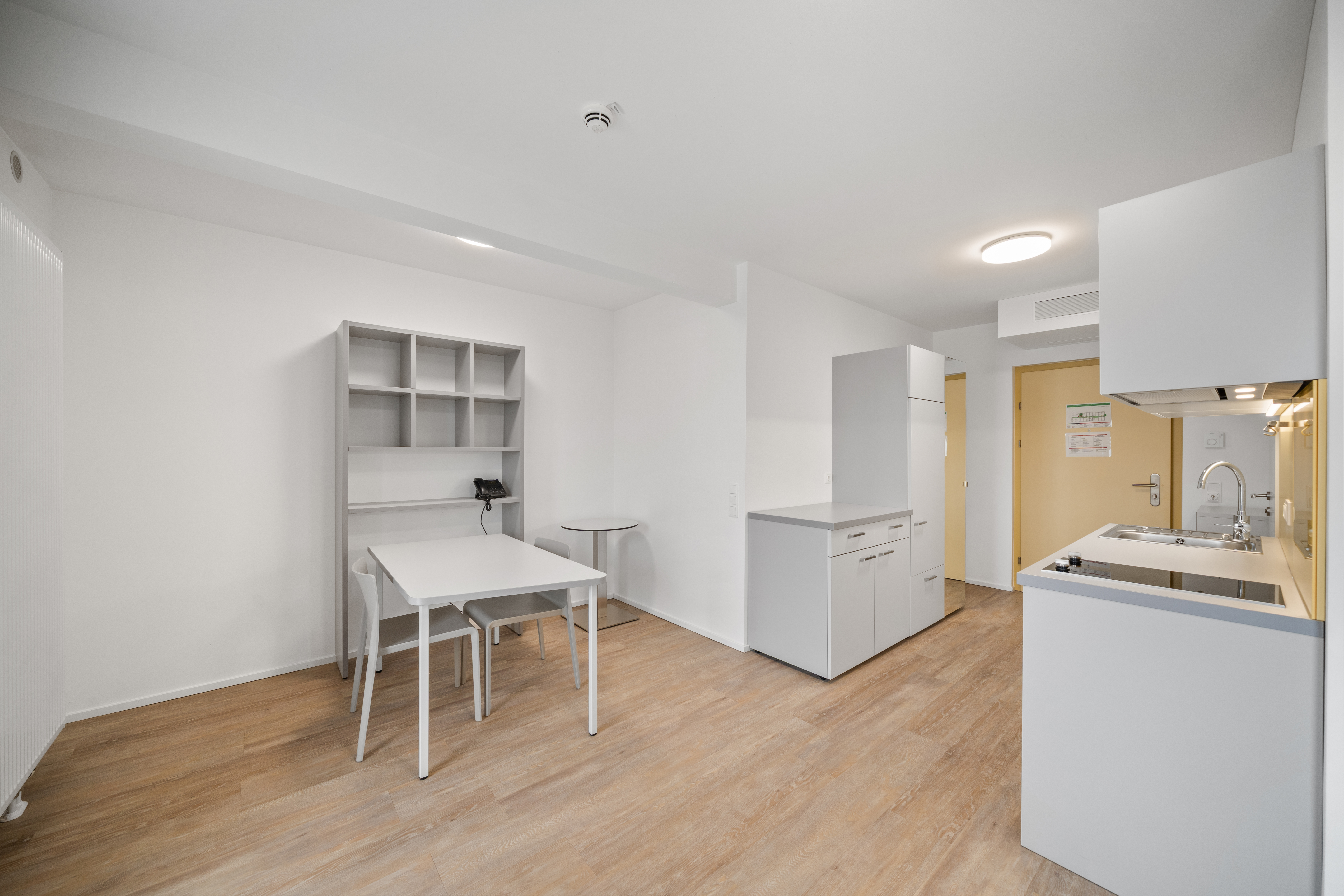 Wohnzimmer mit Arbeitsbereich, weißer Schreibtisch, Regal und offener Küche mit Kücheninsel im möblierten Apartment