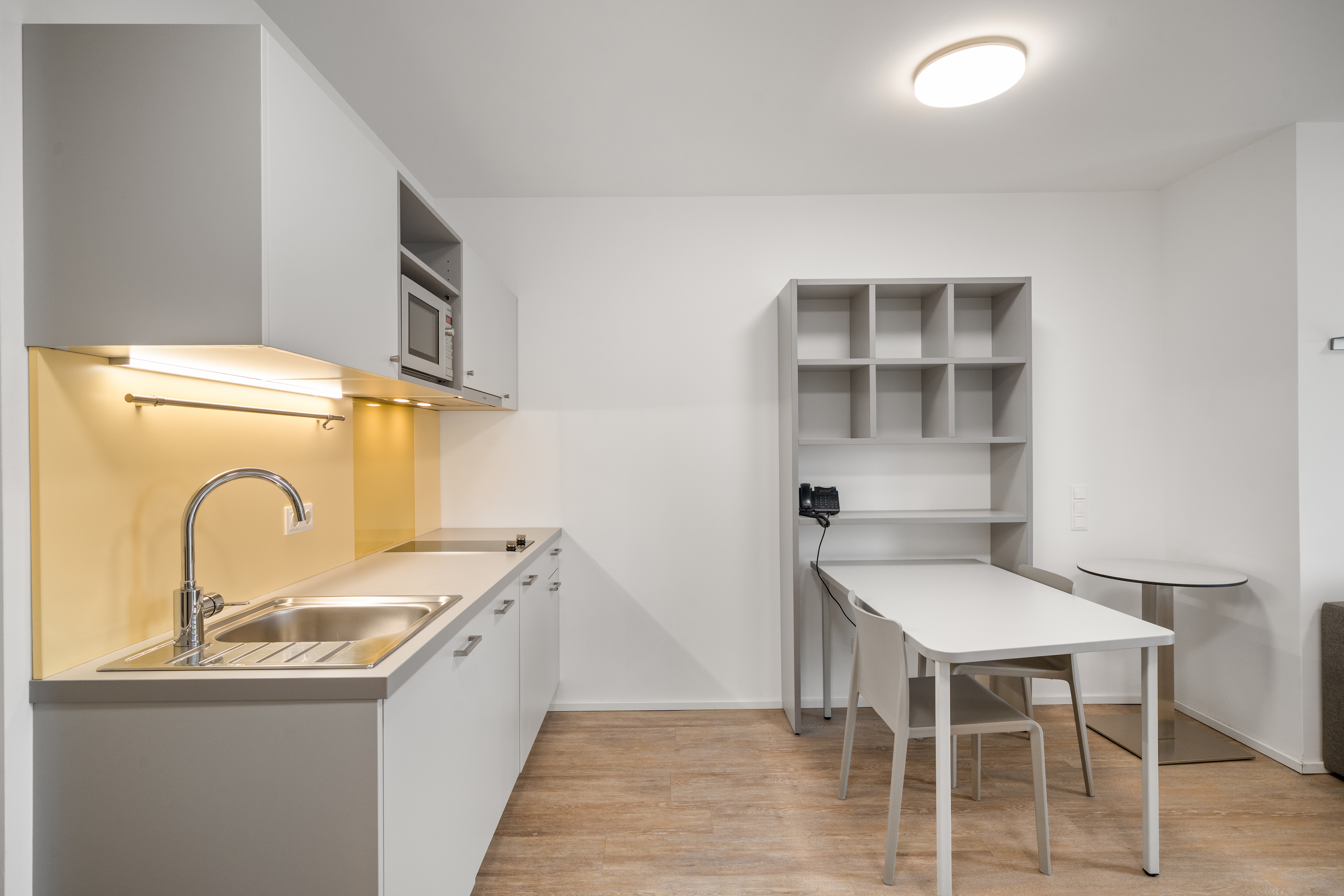 Wohnküche mit weißen Schränken, gelber Rückwand, Esstisch und offenen Regalen im möblierten Apartment Typ M