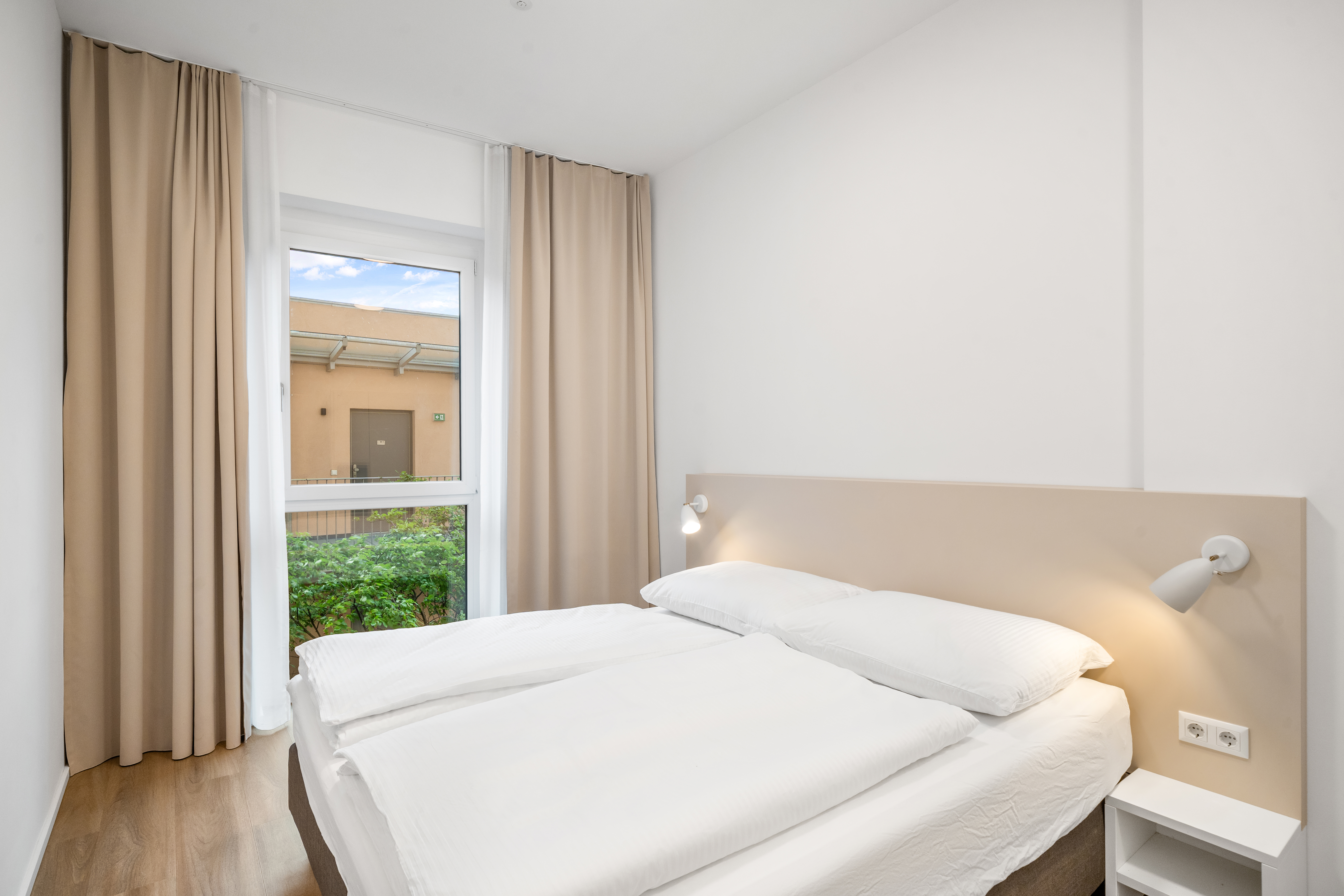 Schlafzimmer mit Doppelbett, weißer Bettwäsche und beigen Vorhängen im Room4Rent Serviced Apartment Typ L