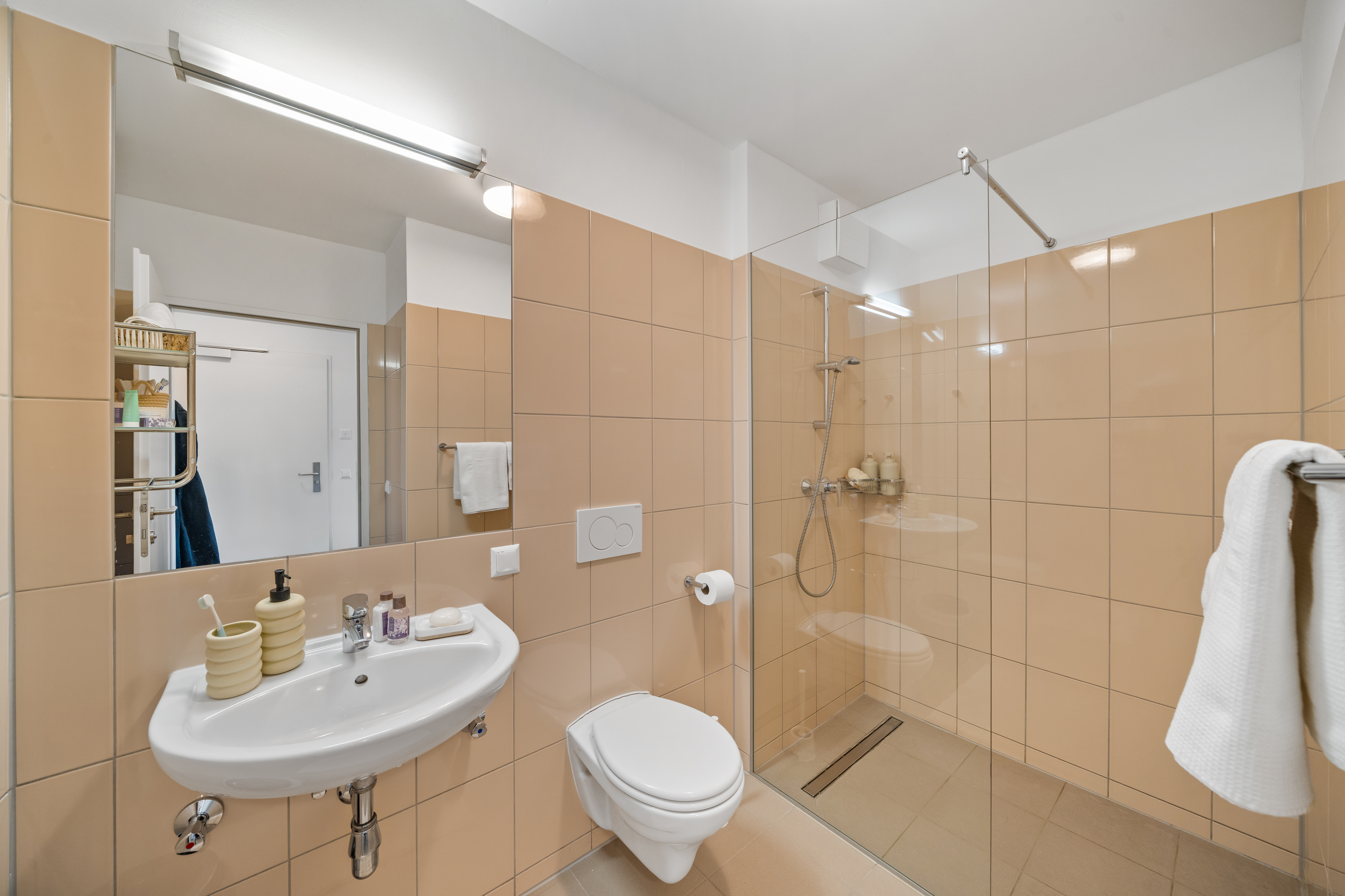 Modernes Badezimmer mit beige Fliesen, weißem Waschbecken, Toilette, begehbarer Dusche mit Glas-Trennwand, Handtuchregal und Badausstattung