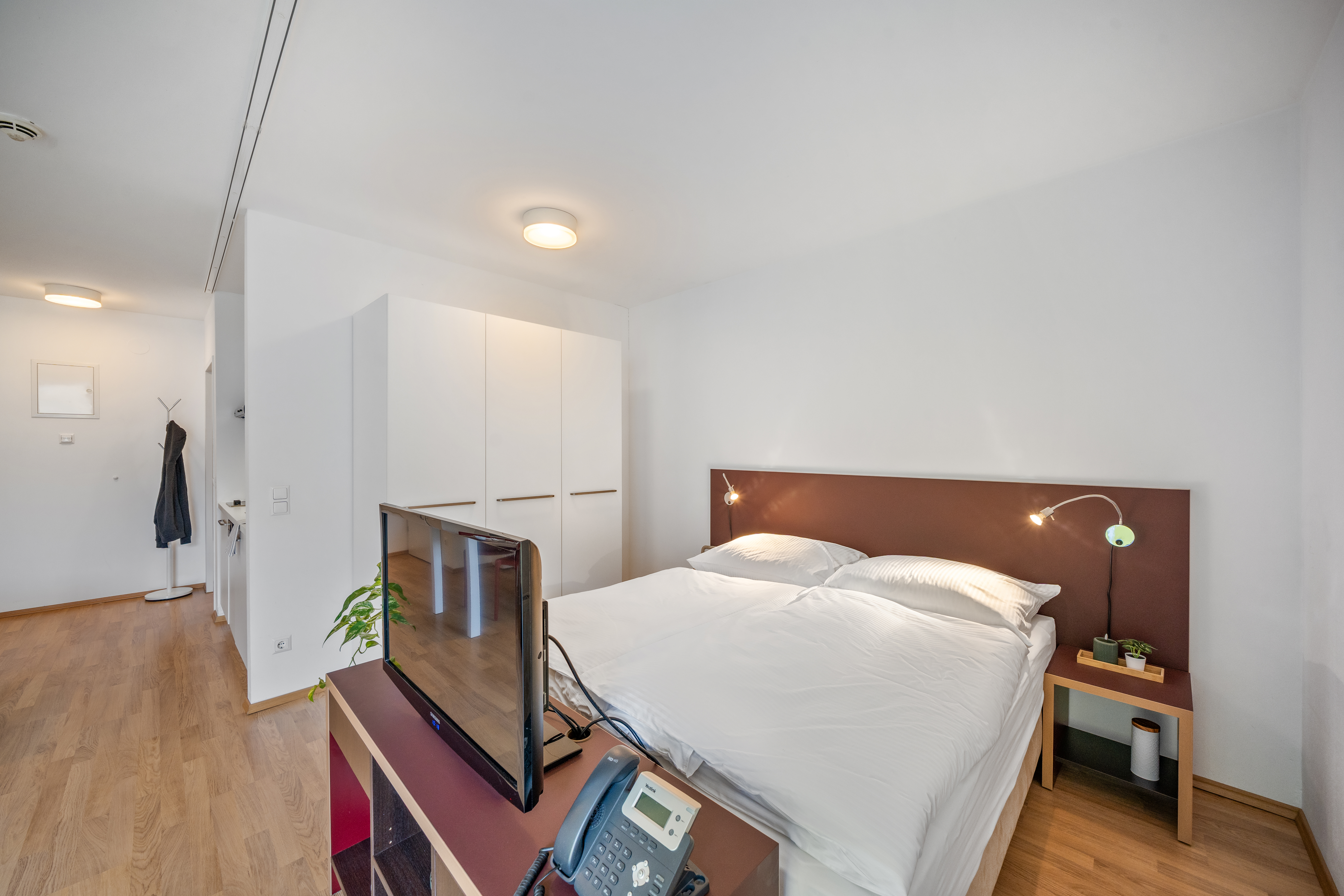 Schlafzimmer mit Doppelbett, braunem Kopfteil, weißem Kleiderschrank und integriertem Arbeitsbereich mit Monitor