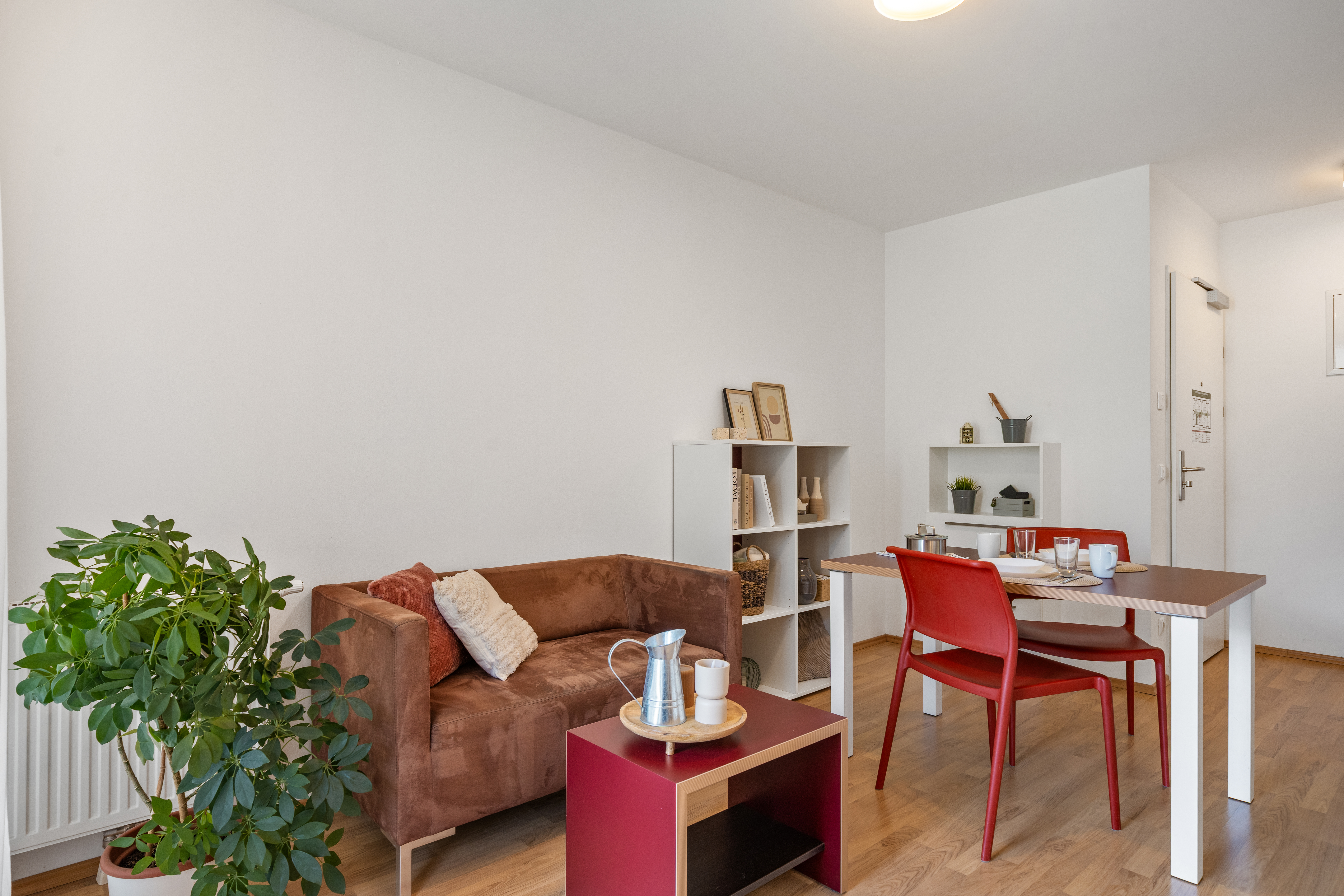 Wohnzimmer mit braunem Sofa, roten Stuhl am Schreibtisch, offenen Regalen und Grünpflanze