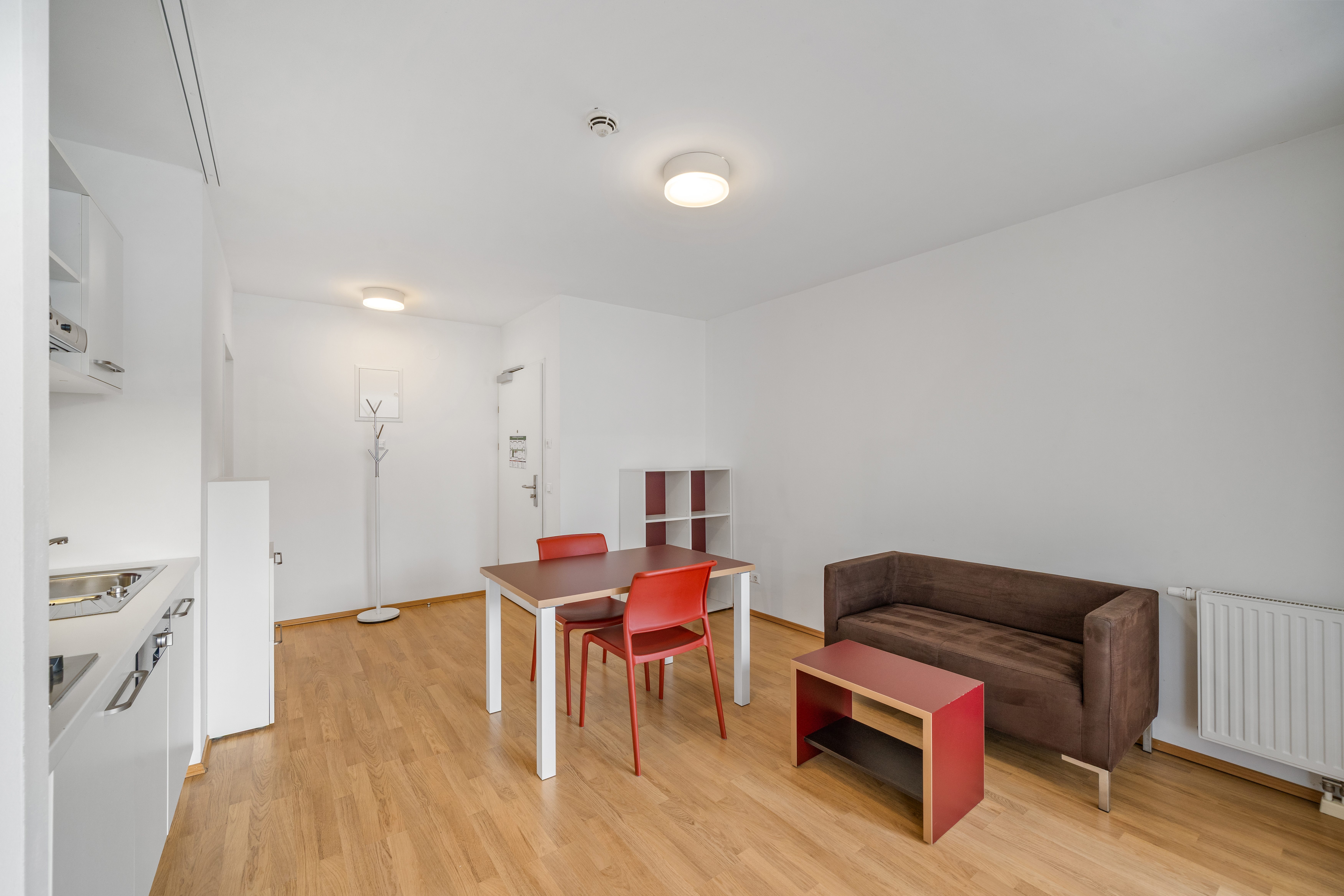 Modernes Wohnzimmer mit Holzfußboden, weiße Küche, roter Esstisch mit Stühlen, braunes Sofa und Wandregal.