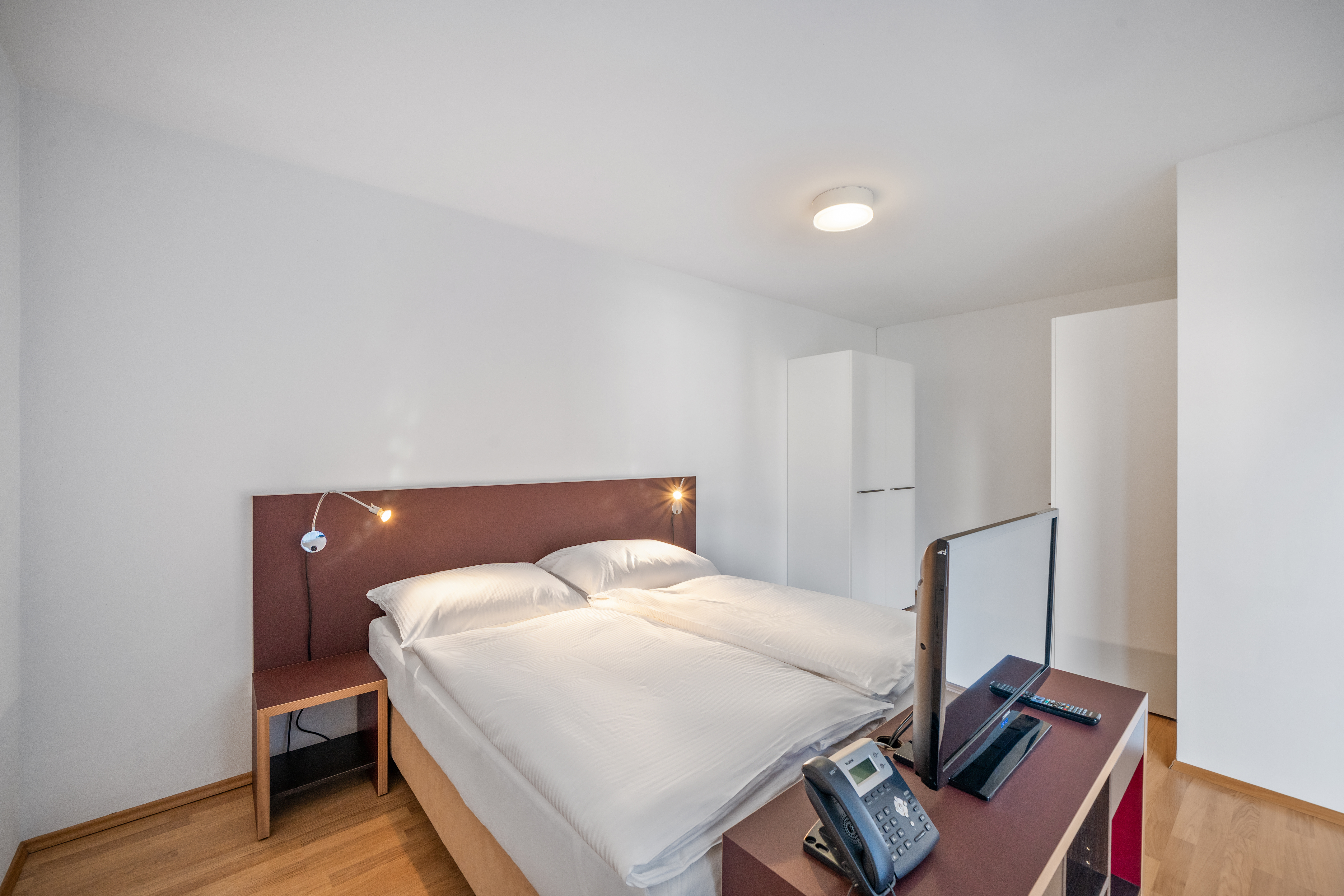 Leeres Schlafzimmer mit Doppelbett, braunem Kopfteil mit Leuchten, Parkettboden und weißen Einbauschränken