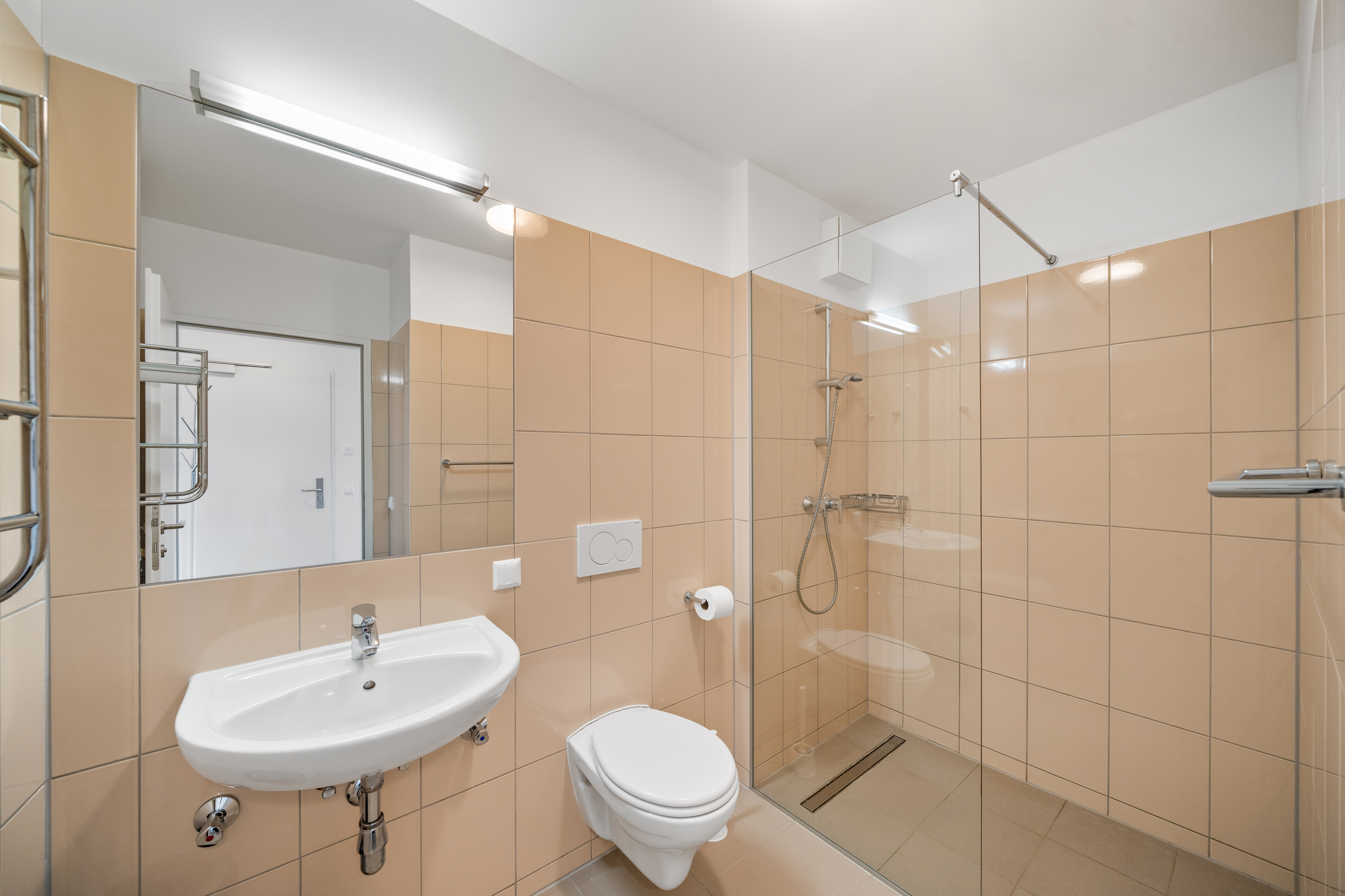Badezimmer mit beigen Wandfliesen, Waschbecken, Spiegel, begehbarer Dusche und Toilettensitz