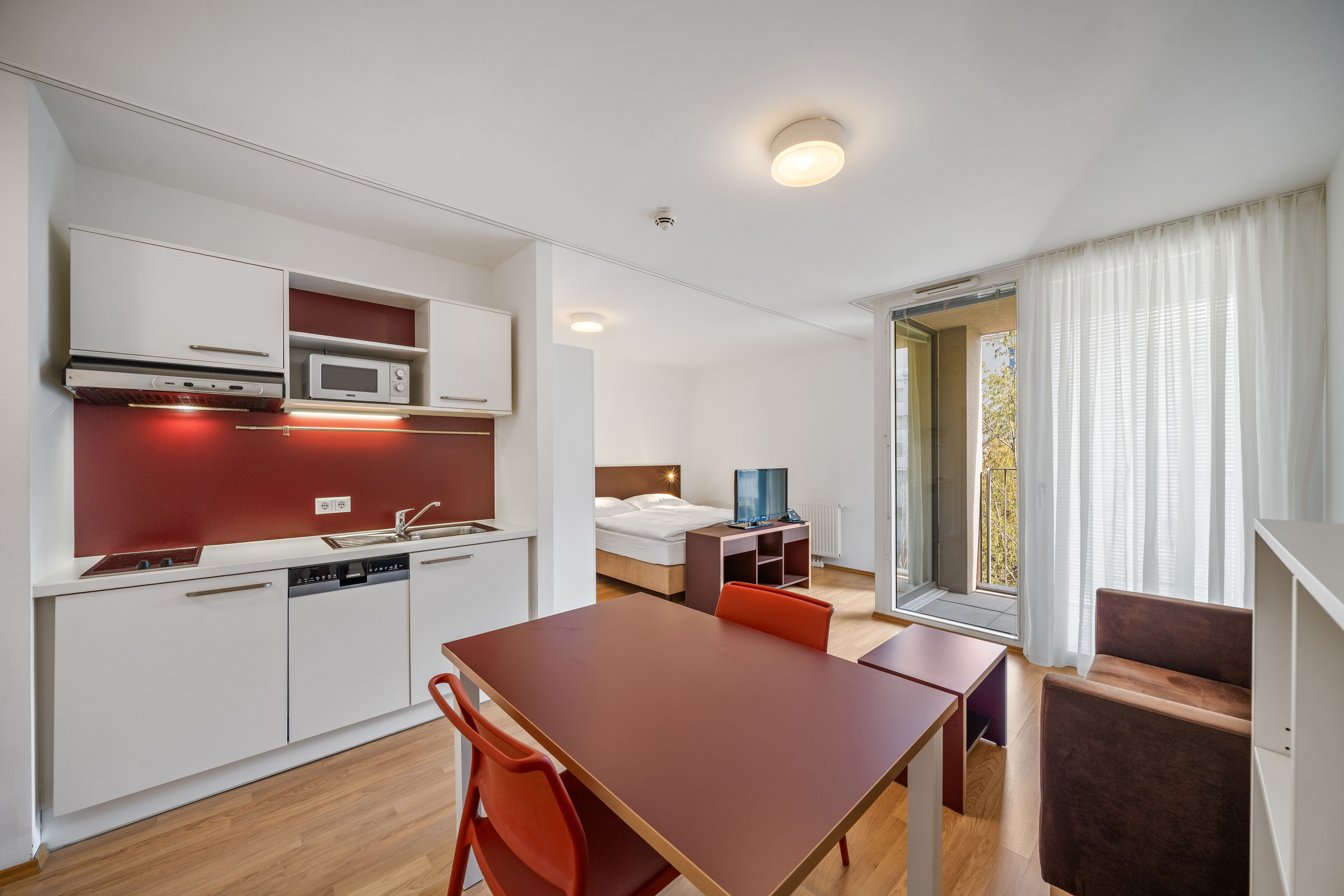 Wohnbereich mit offener Küche, Esstisch und roten Stühlen sowie Schlafbereich mit Balkonzugang im Room4Rent Serviced Apartment am Storchen&shy;grund