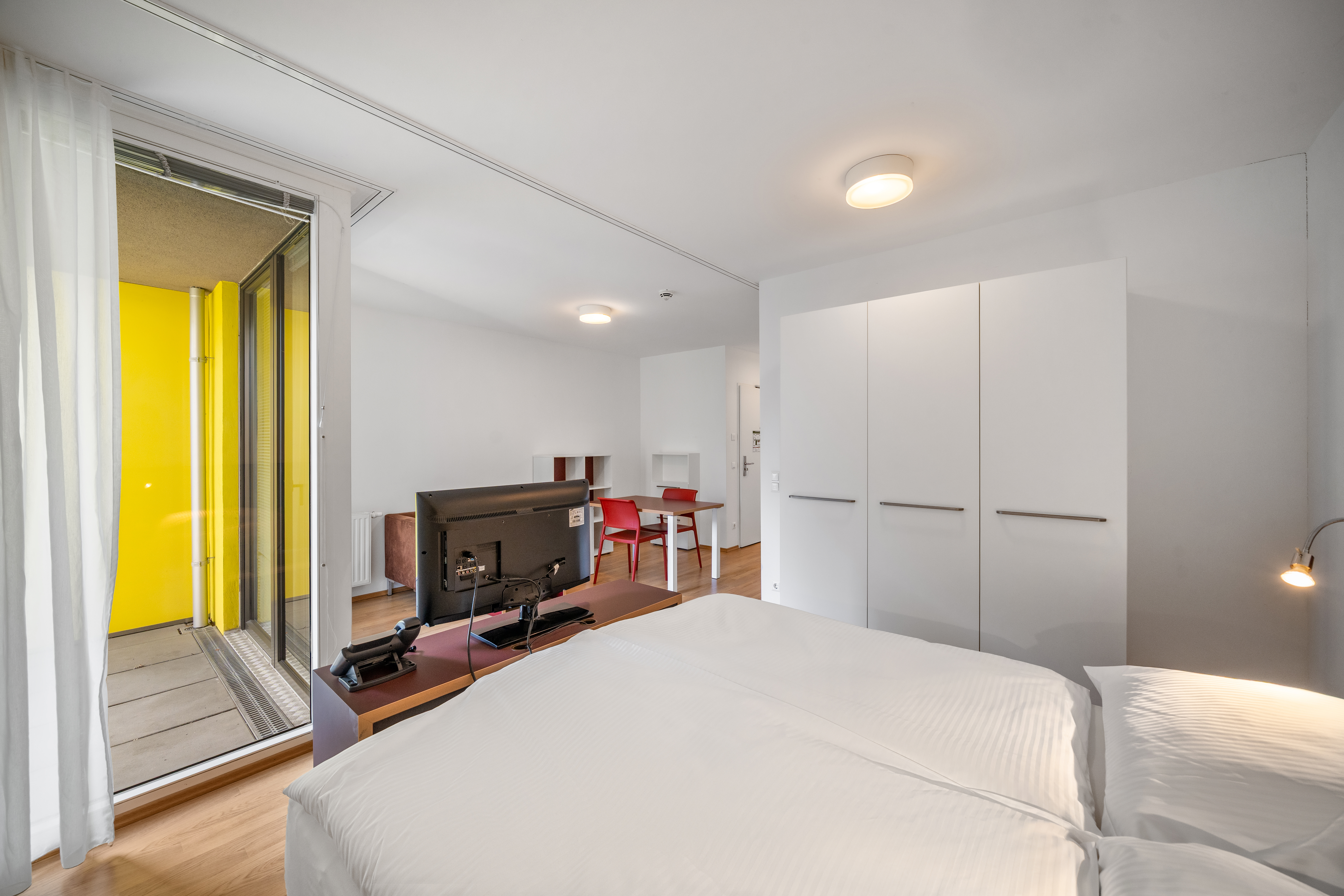 Wohn- und Schlafbereich mit Doppelbett, offener Küche, Esstisch mit roten Stühlen und Balkonzugang im möblierten Room4Rent Apartment am Storchen&shy;grund