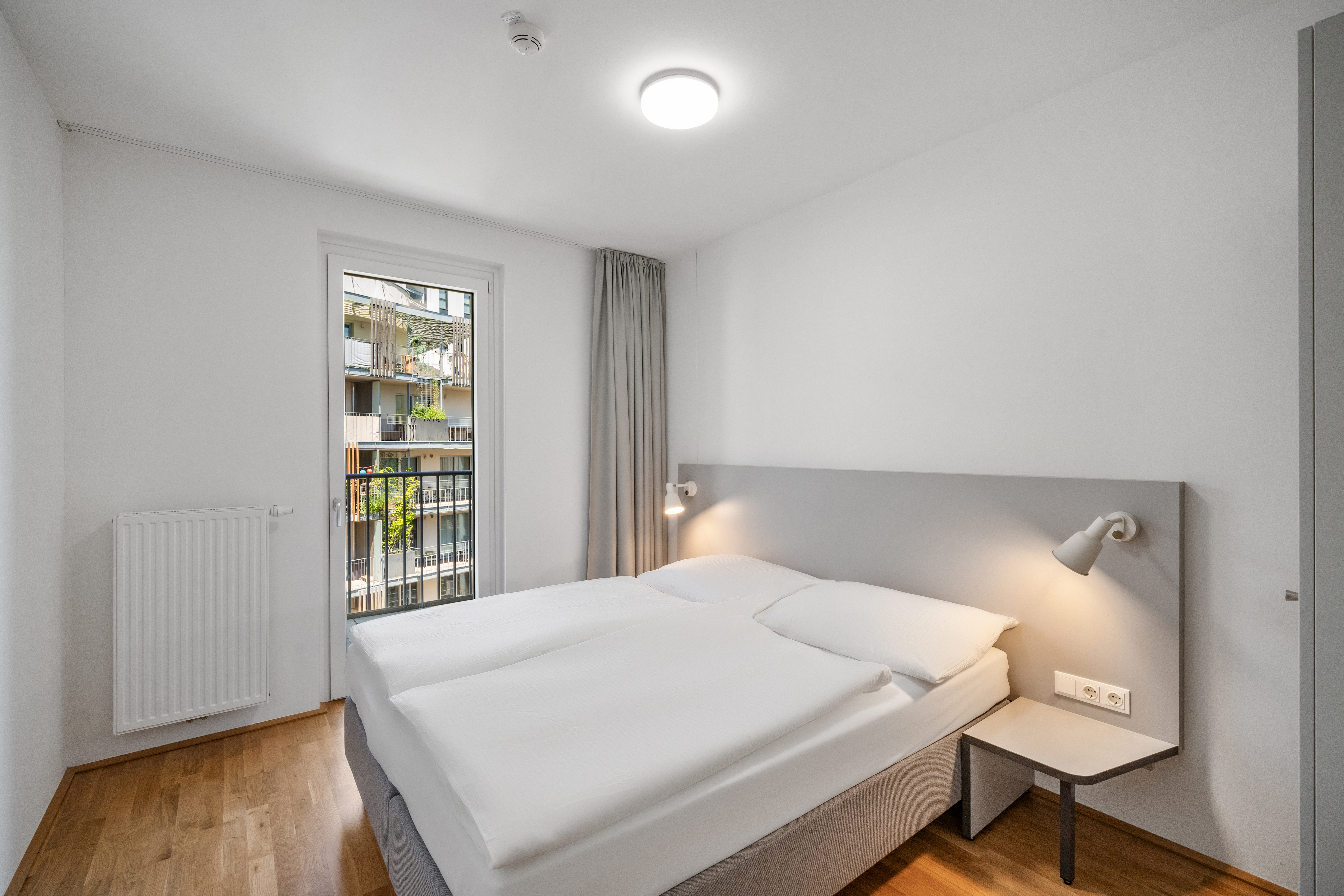 Schlafzimmer im Room4Rent Serviced Apartment mit Doppelbett, grauem Kopfteil, Nachttischen und großem Balkon-Fenster