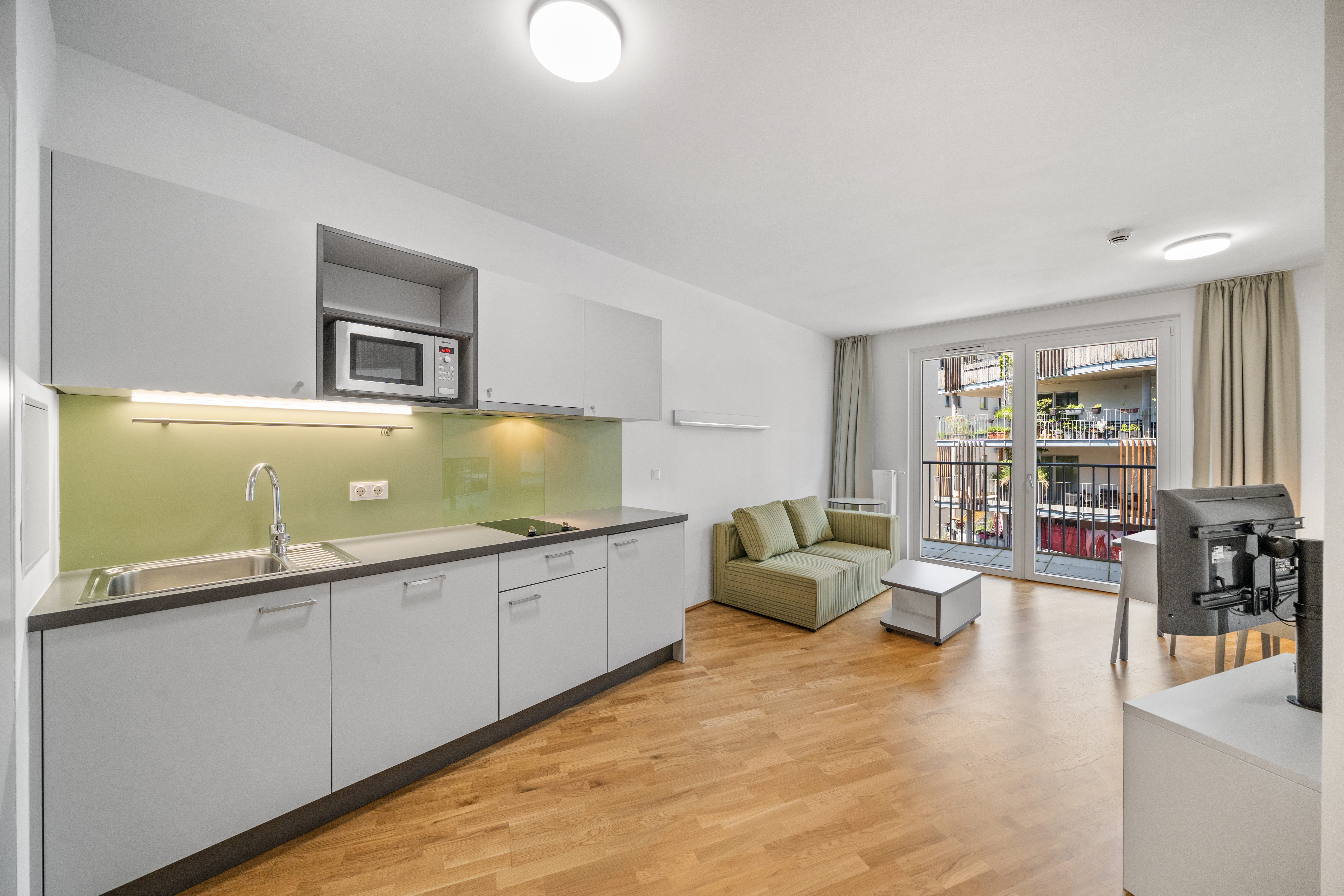 Offener Wohnbereich mit moderner Küche, grünem Sofa und Balkonzugang im Room4Rent möblierten Apartment