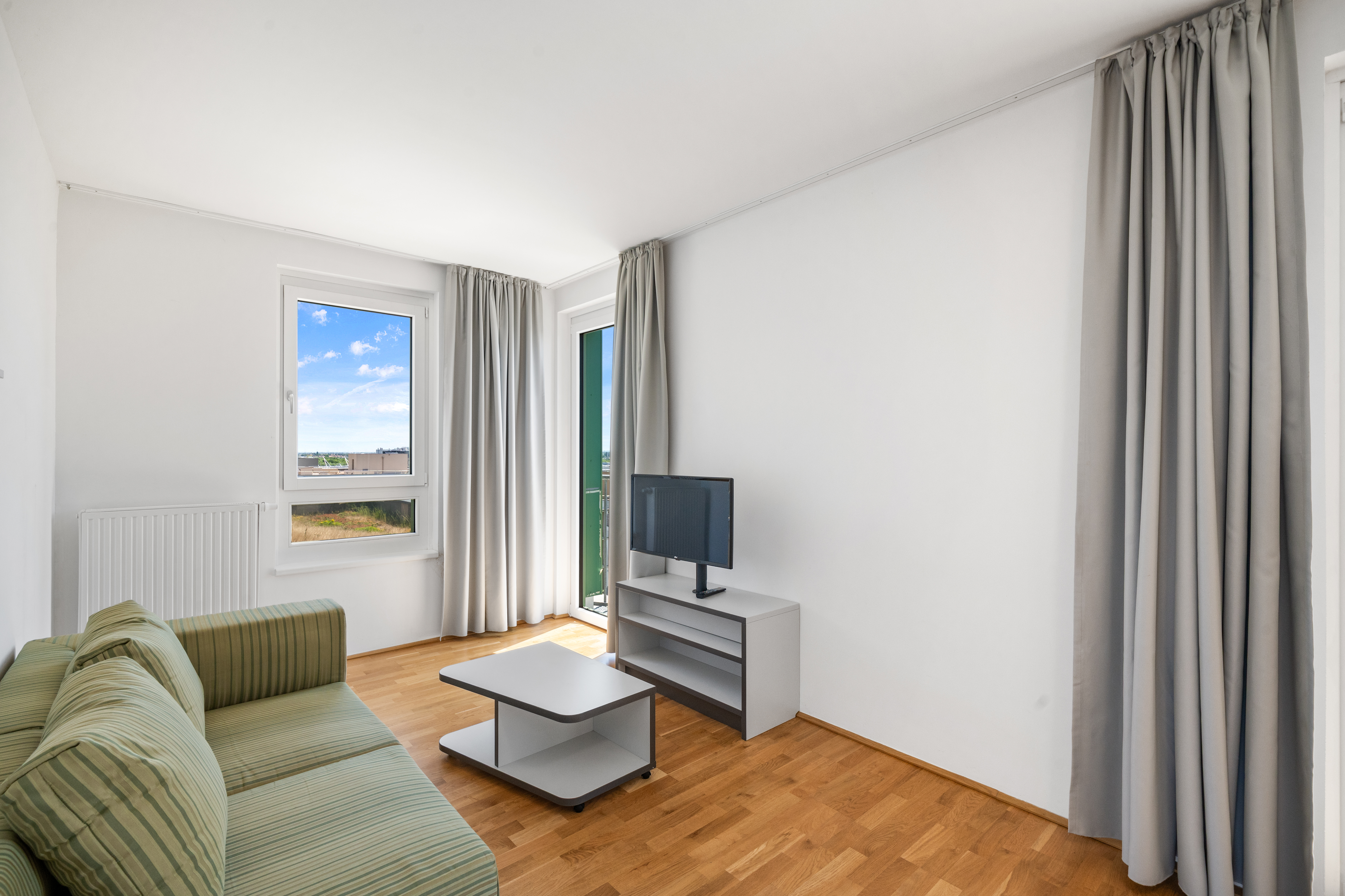 Wohnbereich eines leeren XL-Apartments mit grünem Sofa, Fernseher auf weißem TV-Schrank, Couchtisch und Fenster mit Blick