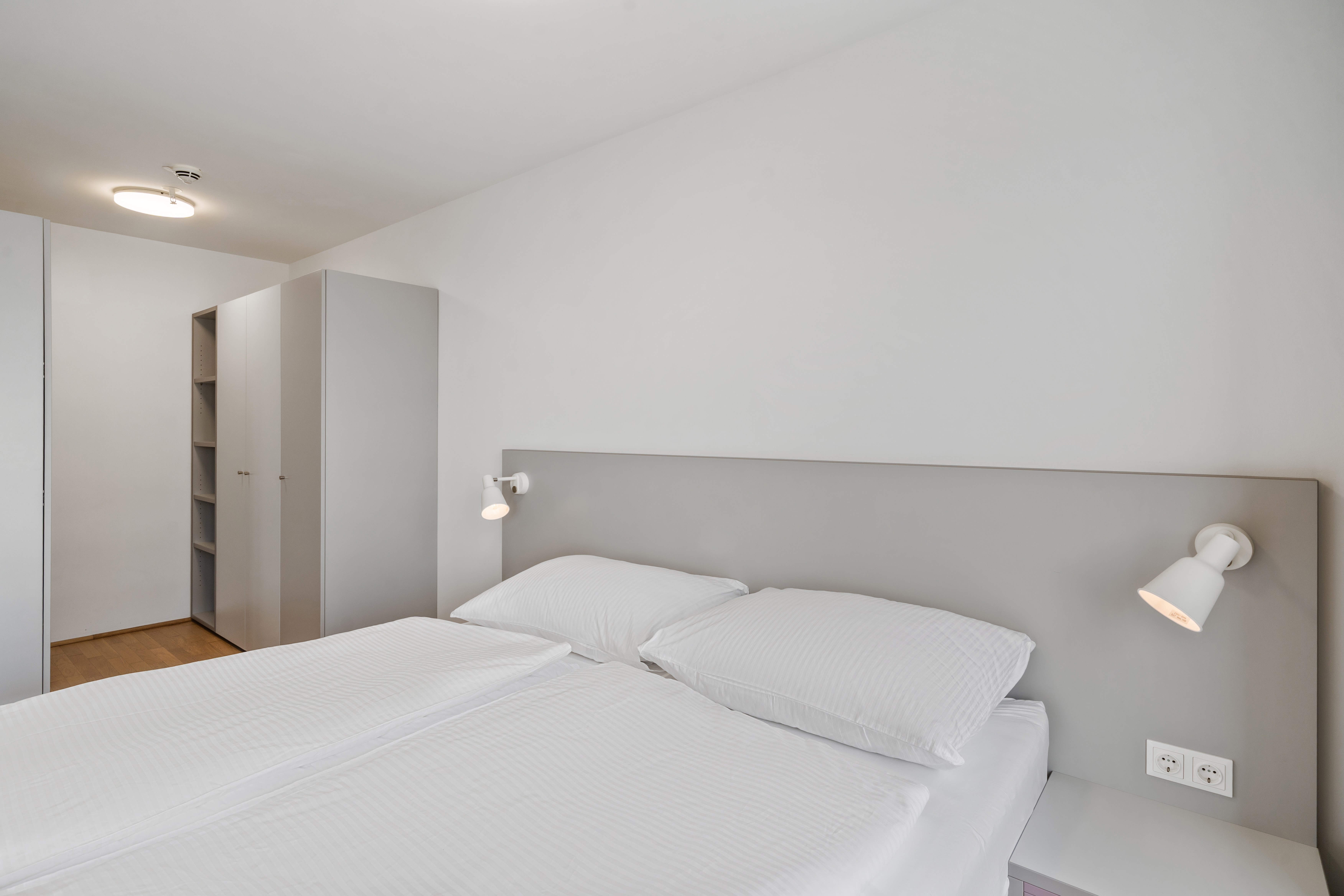 Leeres Schlafzimmer mit Doppelbett, grauem Kopfteil, weißer Bettwäsche, Kissen, zwei Nachttischleuchten und großem Einbauschrank