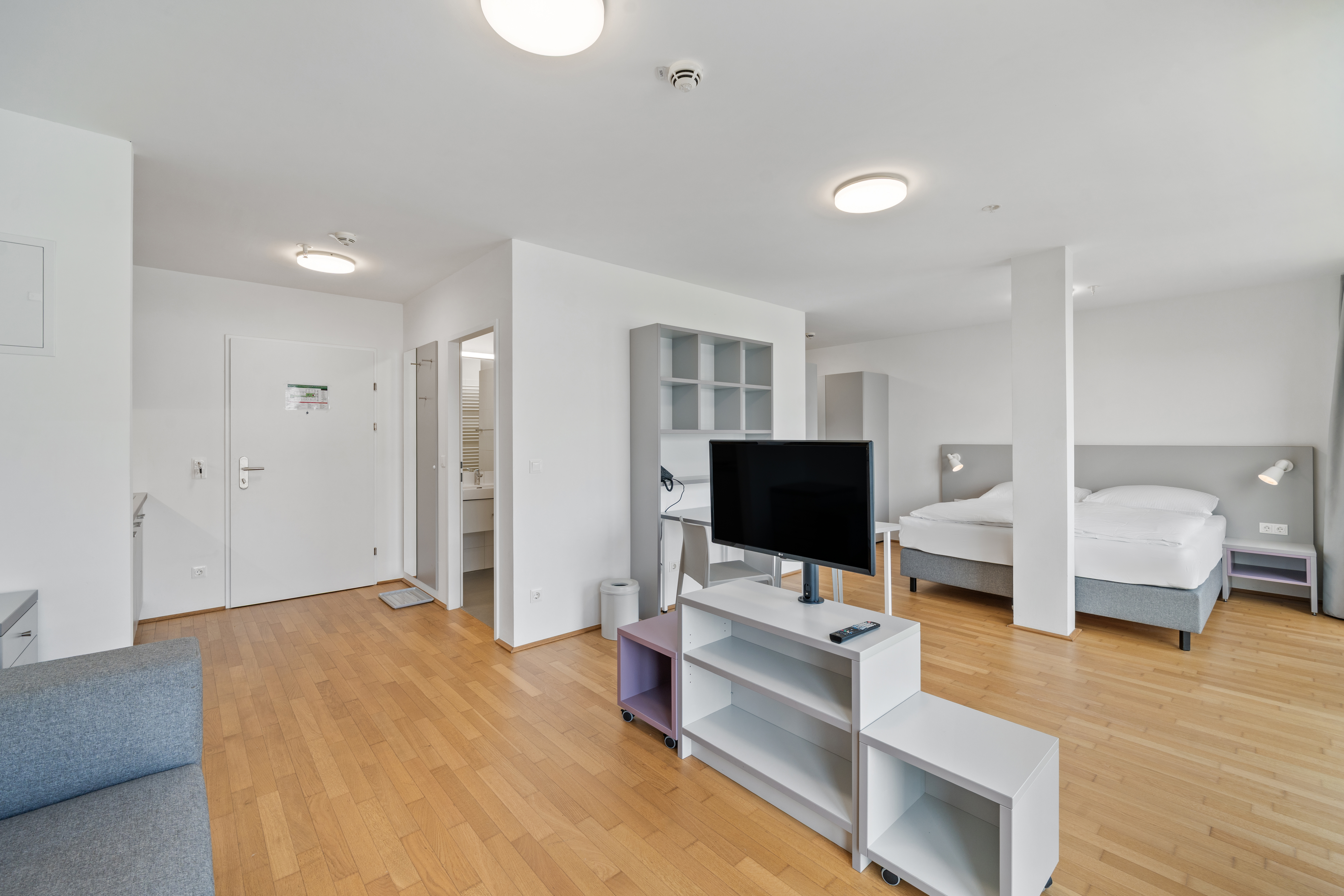 Wohnbereich mit Fernseher auf Regal, Betten im Hintergrund und Holzparkett