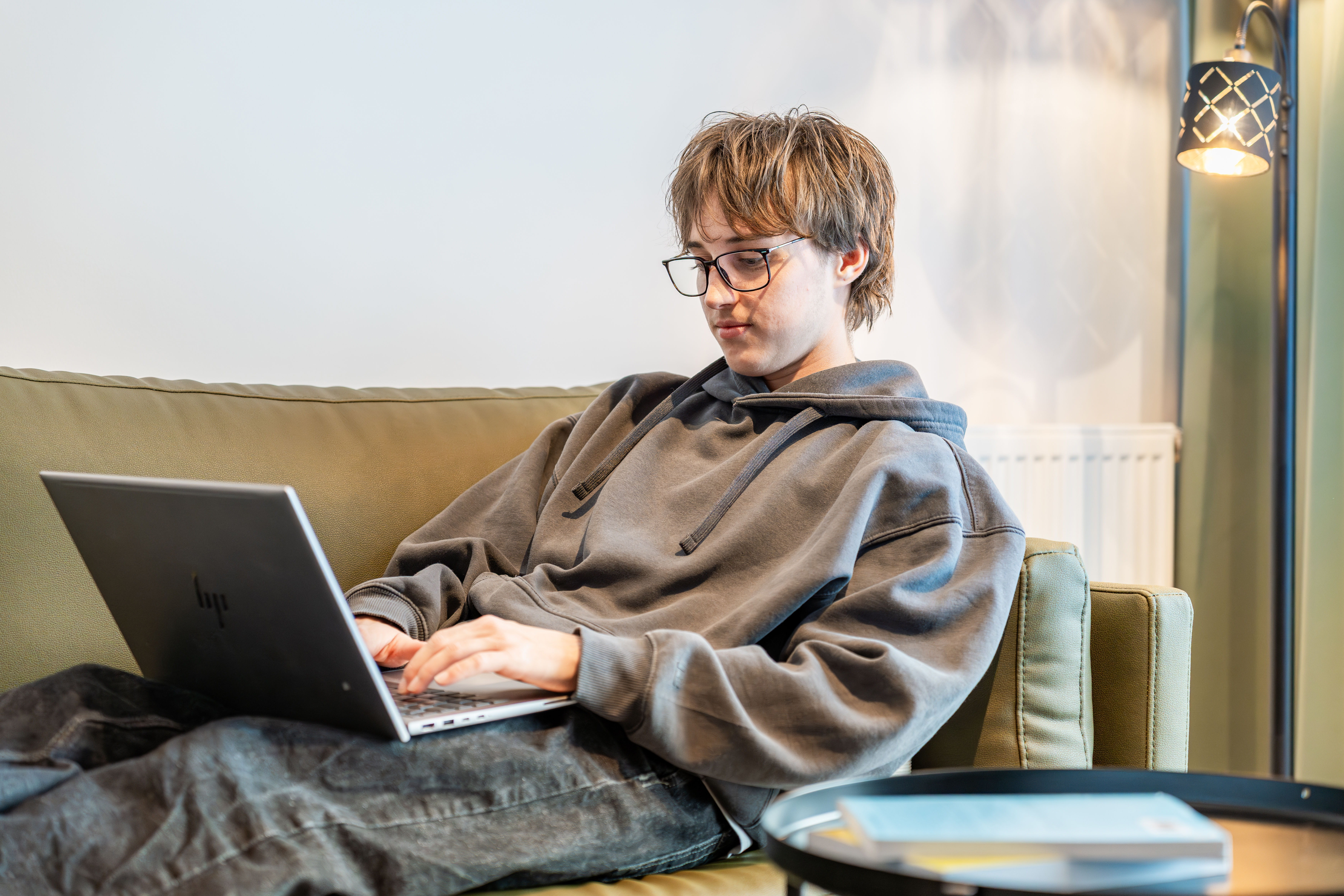Ein junger Student in grauem Pullover arbeitet mit ausgestreckten Beinen auf seiner Couch an seinem Laptop.